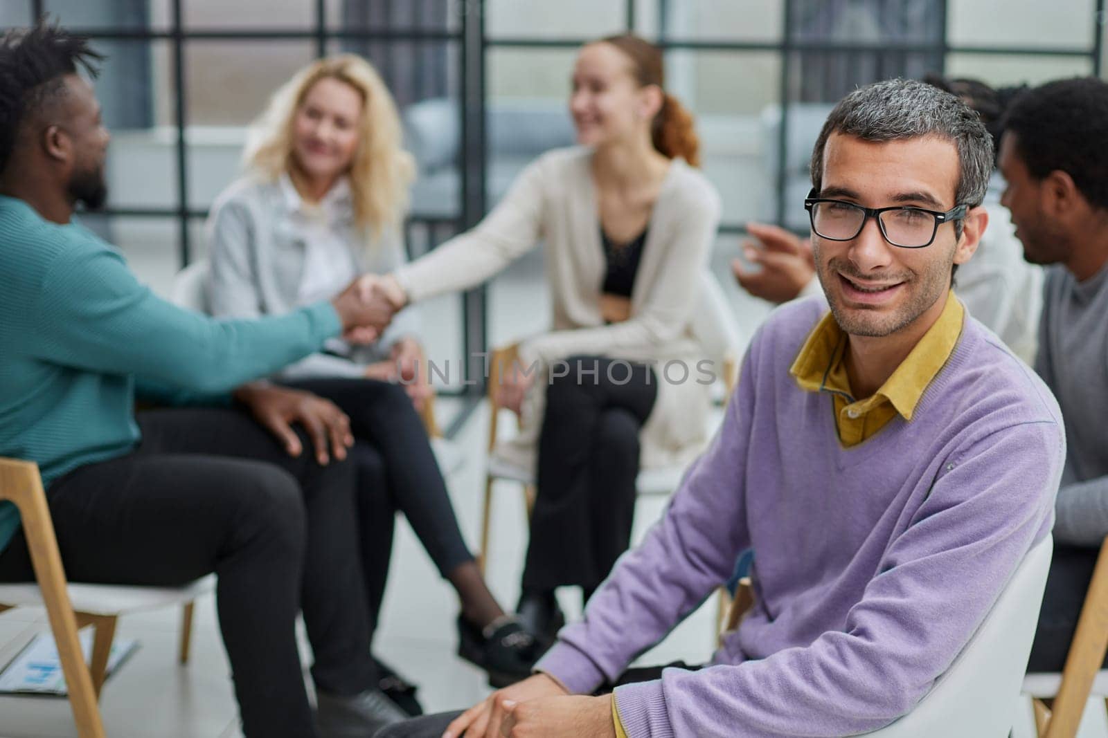A happy young businessman, in a purple sweater, sits against the background of his colleagues in a modern office