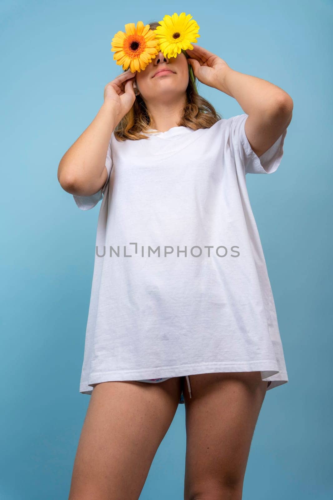 portrait of a funny teenage girl with yellow gerbera flowers on her eyes, standing on a bright blue background in a white T-shirt and shorts. the girl holds gerbera flowers over her eyes. A playful girl covers her eyes with yellow flowers. vertical photo