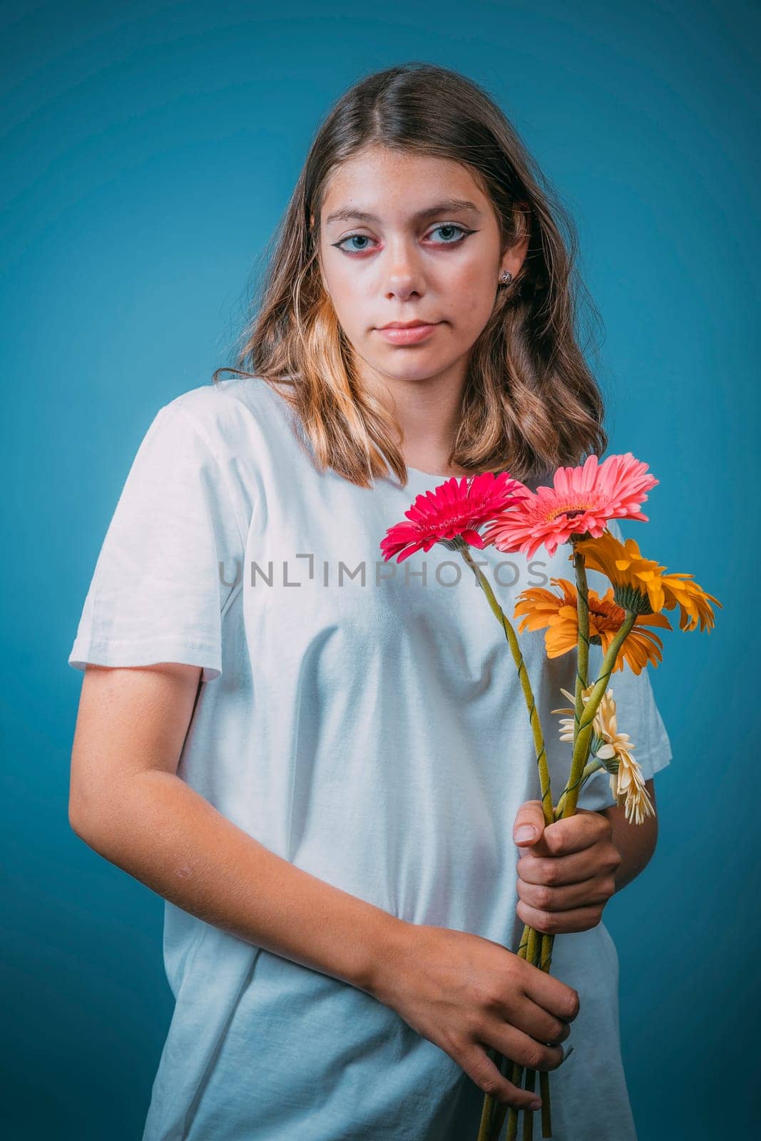 portrait of a pretty Caucasian girl with gerbera flowers on a blue background. A teenage girl in a white T-shirt with brown hair holds flowers and looks at the camera