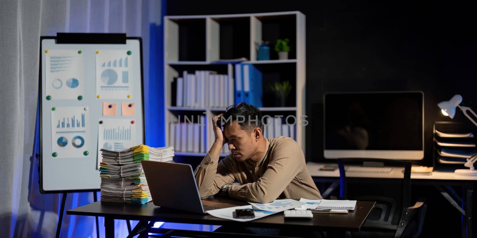 Overworked young Asian office employee working on laptop computer overtime in office at night.