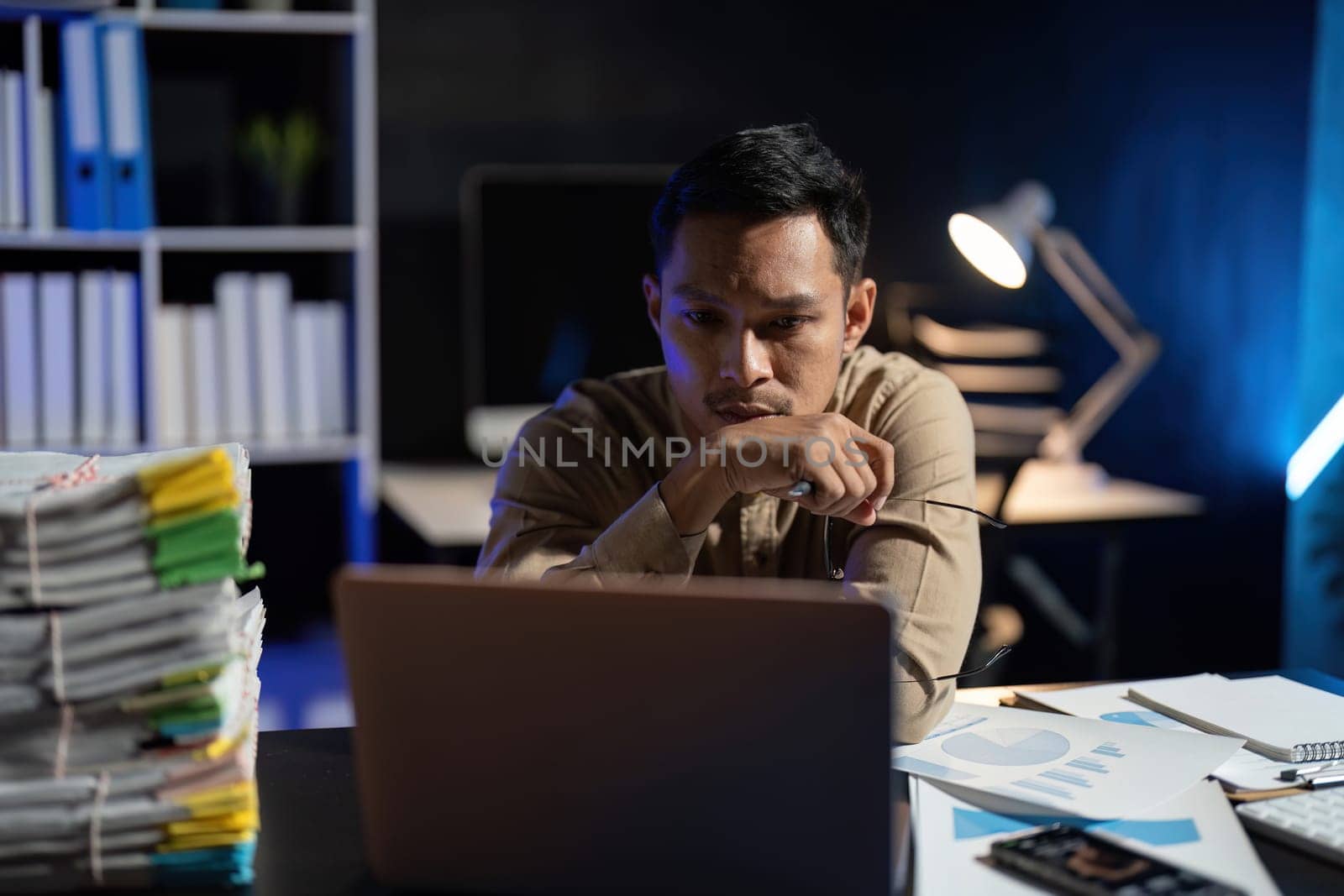 Overworked young Asian office employee working on laptop computer overtime in office at night.