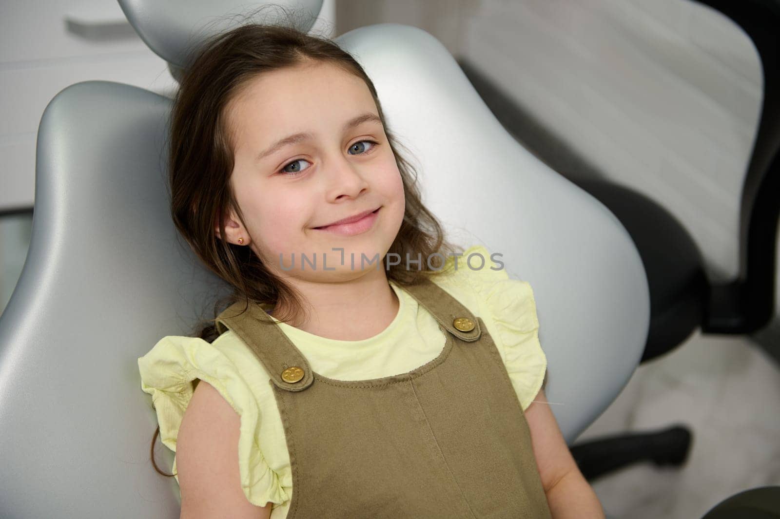 Beautiful kid girl in dentist chair, smiles looking at camera while an appointment in dentistry clinic. Pediatric dental practice. Prophylactic examination of baby teeth to early prevention caries