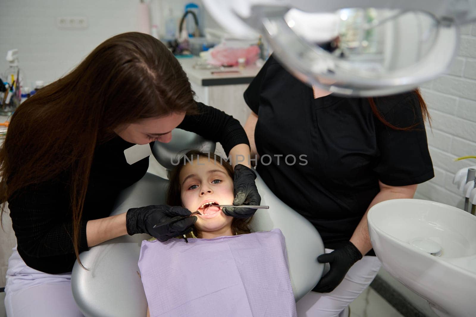 Caucasian adorable little girl with open mouth, sitting in dentist chair, getting teeth examination in pediatric dentistry clinic. Woman dental hygienist using dental mirror checking kid's oral cavity
