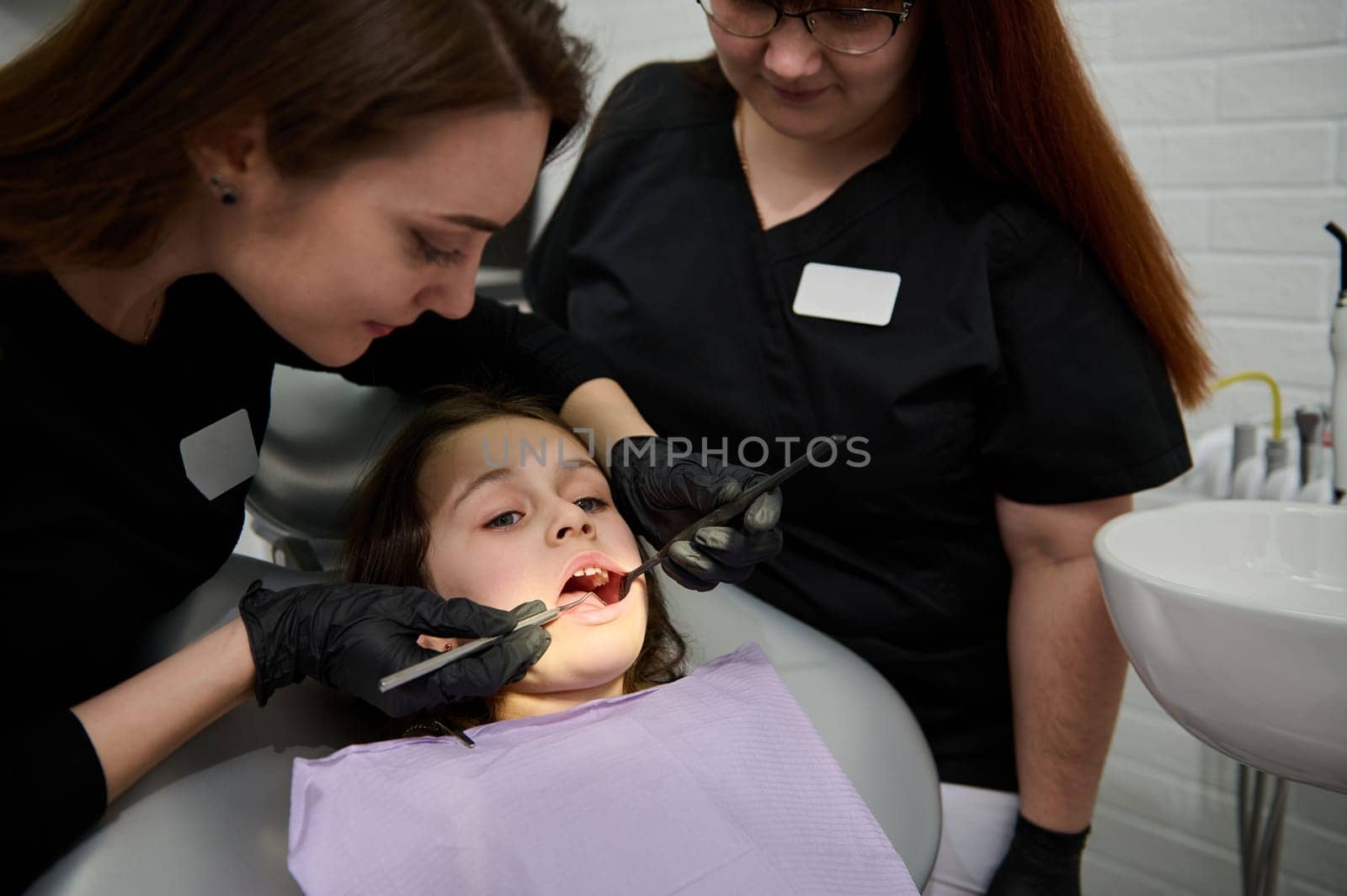 Portrait of beautiful child 6 years old, adorable little girl with open mouth, being examined by a dental hygienist, orthodontist while sitting in dentist chair in pediatric dentistry clinic. Close-up