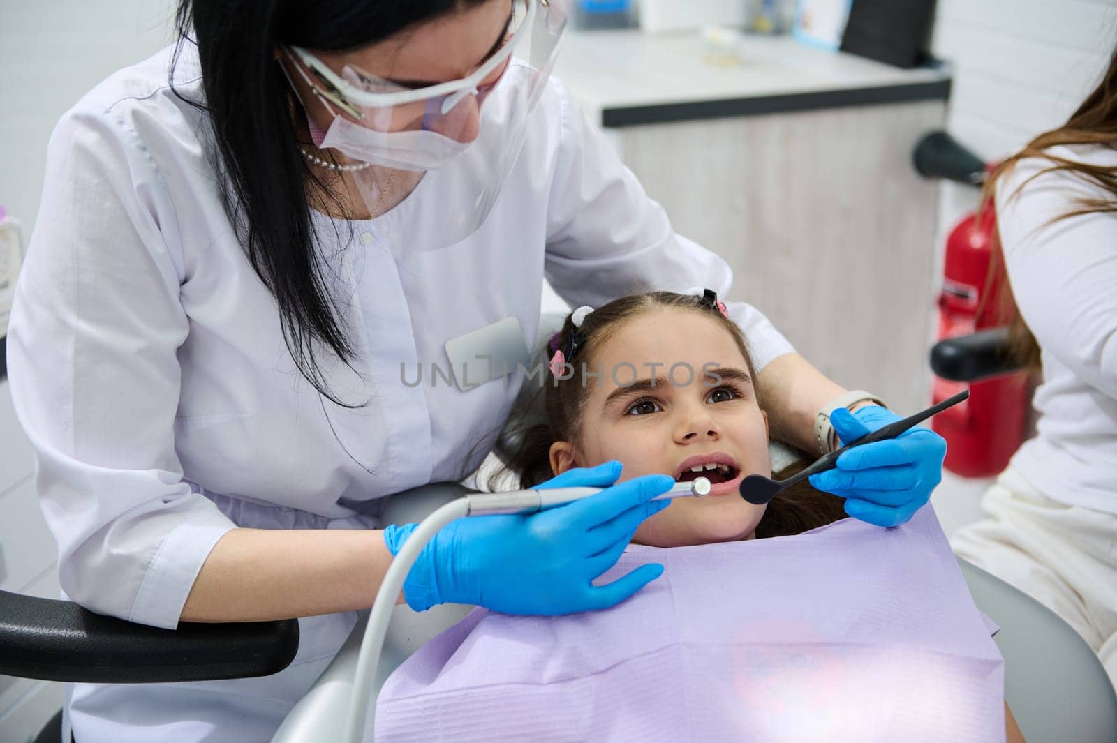 Female dentist doctor in medical protective eyewear, holding dental tools, performing dental treatment and cleaning to a little child girl in dentist's chair, curing caries in dentistry clinic