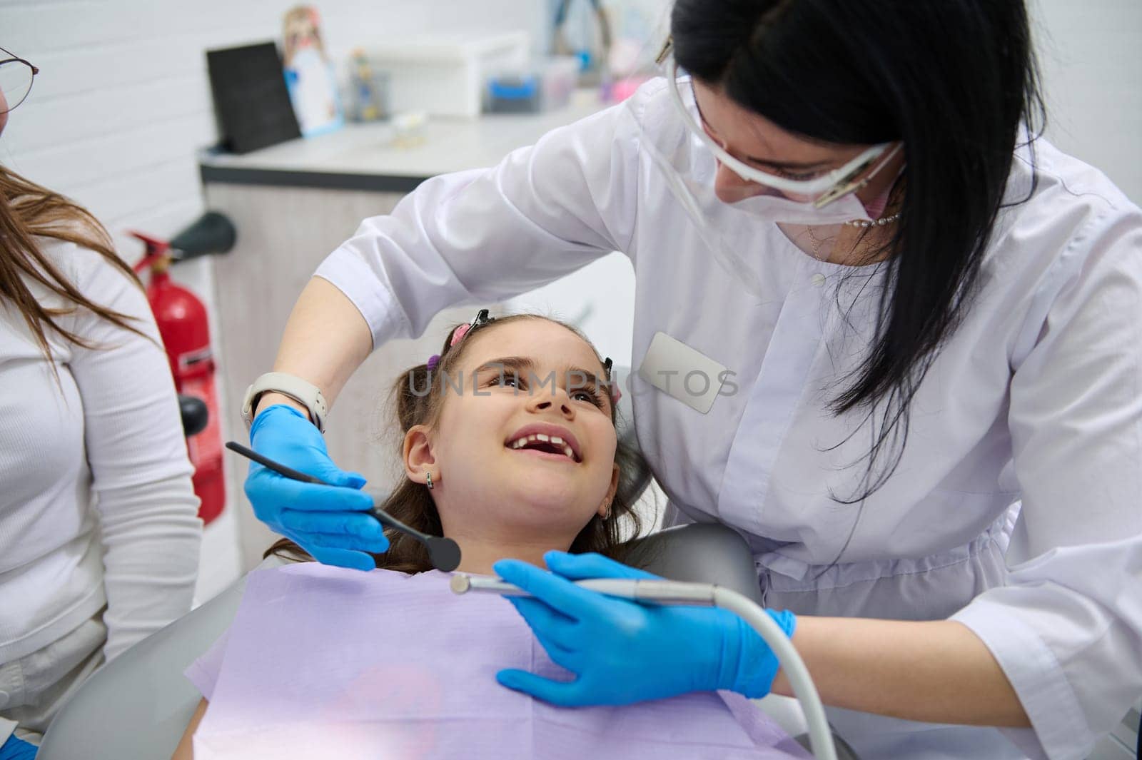 Happy little child girl, smiling to her dentist doctor while dental appointment in dentistry clinic. Pleasant female dental hygienist, orthodontist doing teeth and mouth cavity check up. Dental health