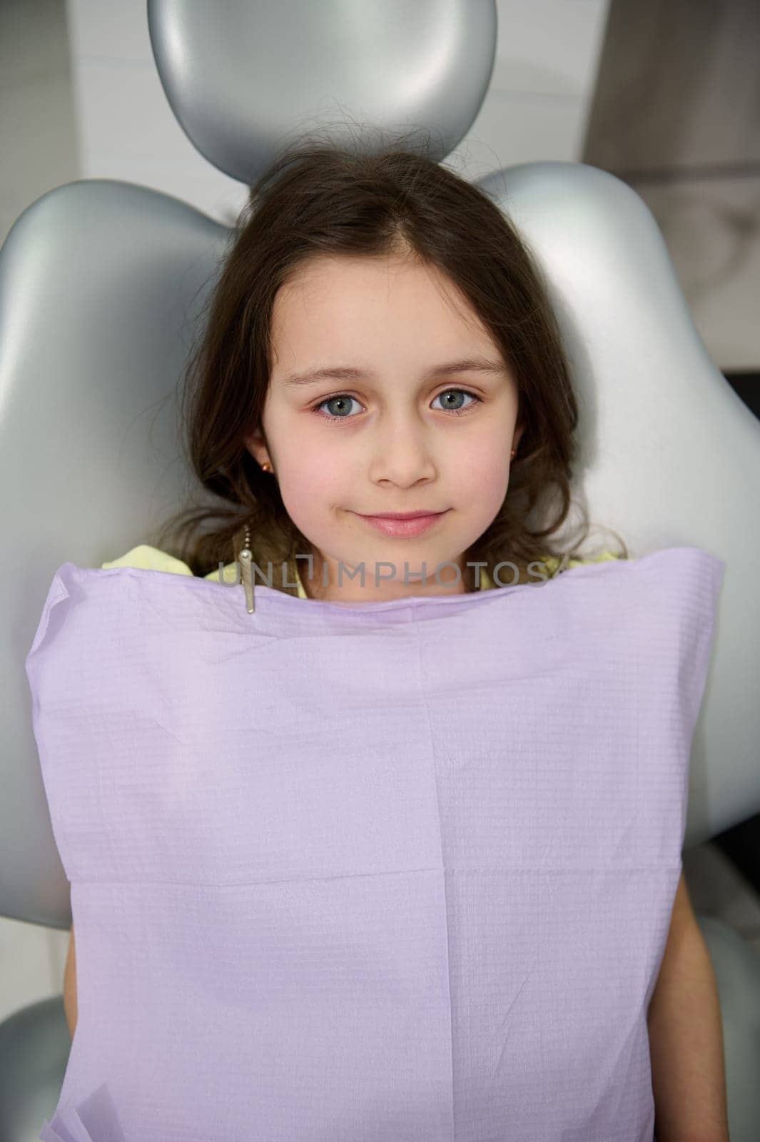 Top view. Beautiful Caucasian child, lovely little girl in dentist's chair, smiling looking at camera, ready to get a dental treatment or regular prophylactic checkup of baby teeth in dentistry clinic
