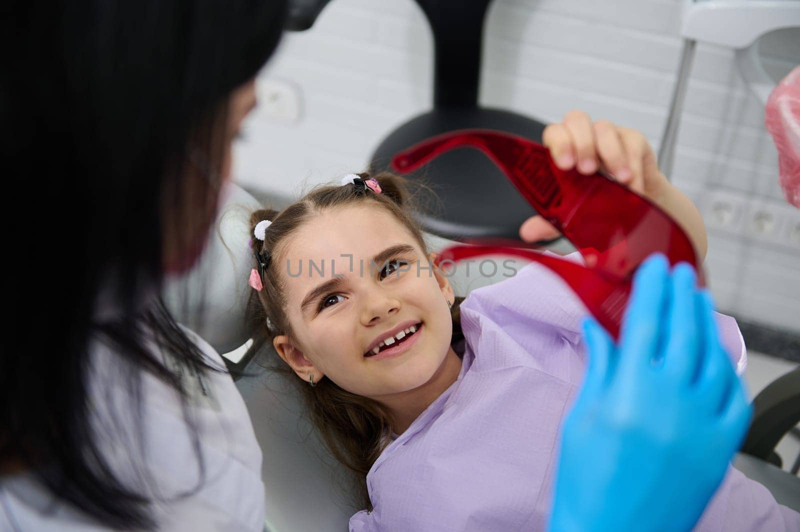 Smiling kid girl visiting dentist, curing tooth, putting braces in modern pediatric dentistry clinic. Female dentist, dental hygienist treating caries to little child patient. Oral health and hygiene