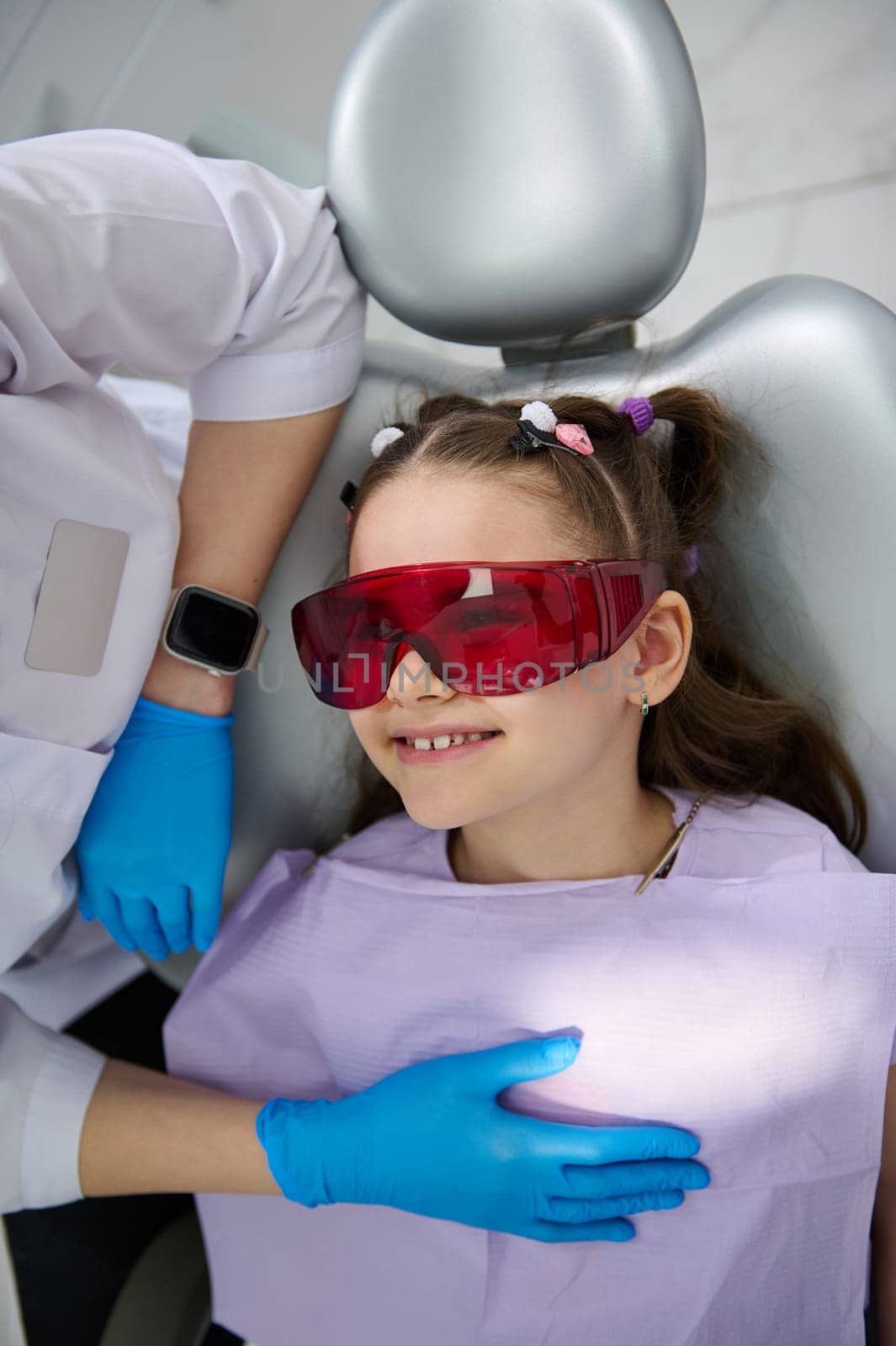 Close-up top view of a smiling happy little child girl, patient in UV goggles, visiting dentist in pediatric dentistry clinic. Dental health and oral hygiene. The concept of baby teeth treatment