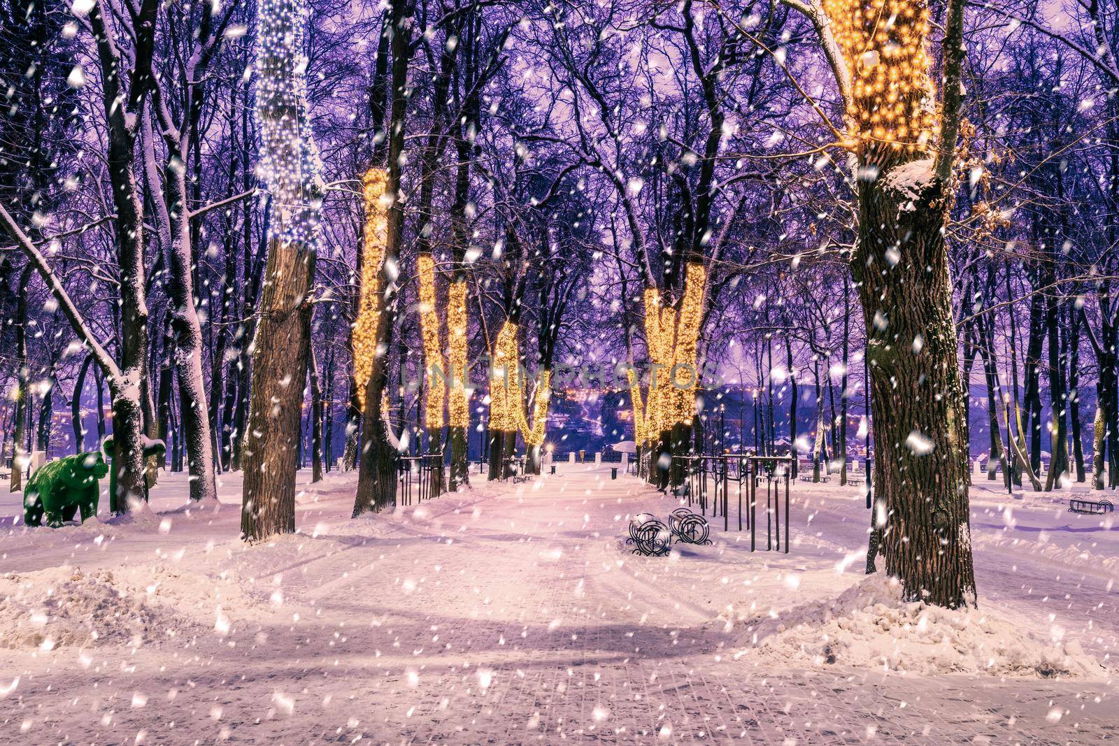 Snowfall in a winter park at night with christmas decorations, lights, pavement covered with snow and trees with garlands.