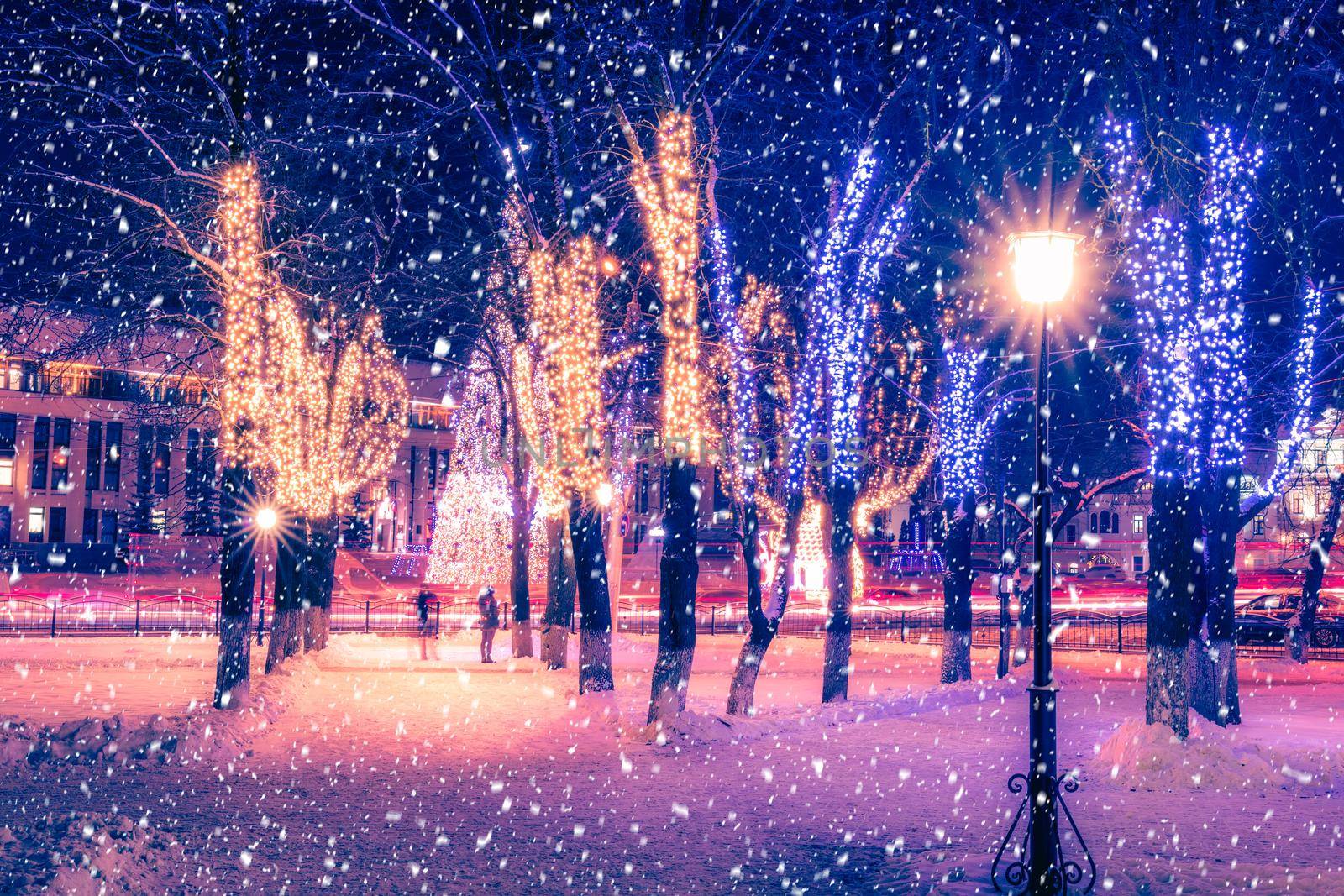 Snowfall in a winter park at night with christmas decorations, lights, pavement covered with snow and trees with garlands.