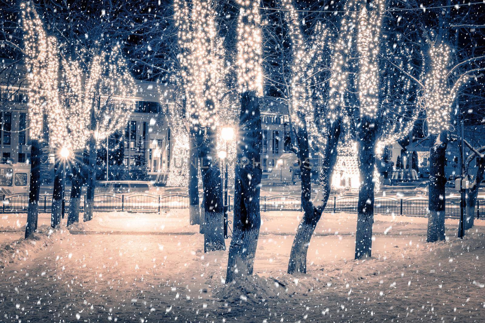 Snowfall in a winter park at night with christmas decorations, lights, pavement covered with snow and trees with garlands.