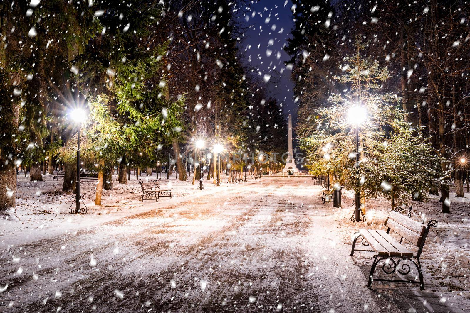 Snowfall in a winter park at night with christmas decorations, lights and  pavement covered with snow. Falling snow.