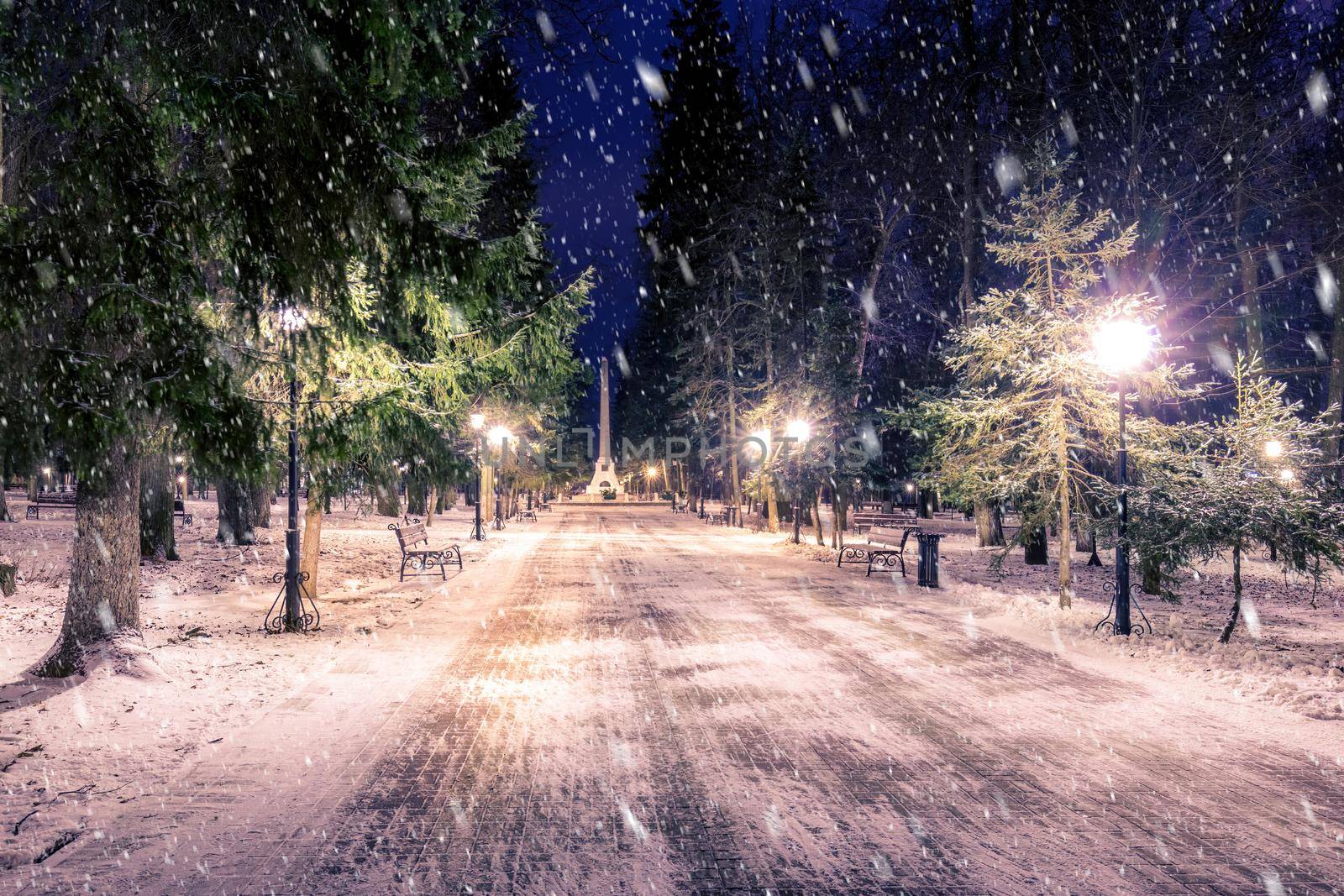 Snowfall in a winter park at night with christmas decorations, lights and  pavement covered with snow. Falling snow.