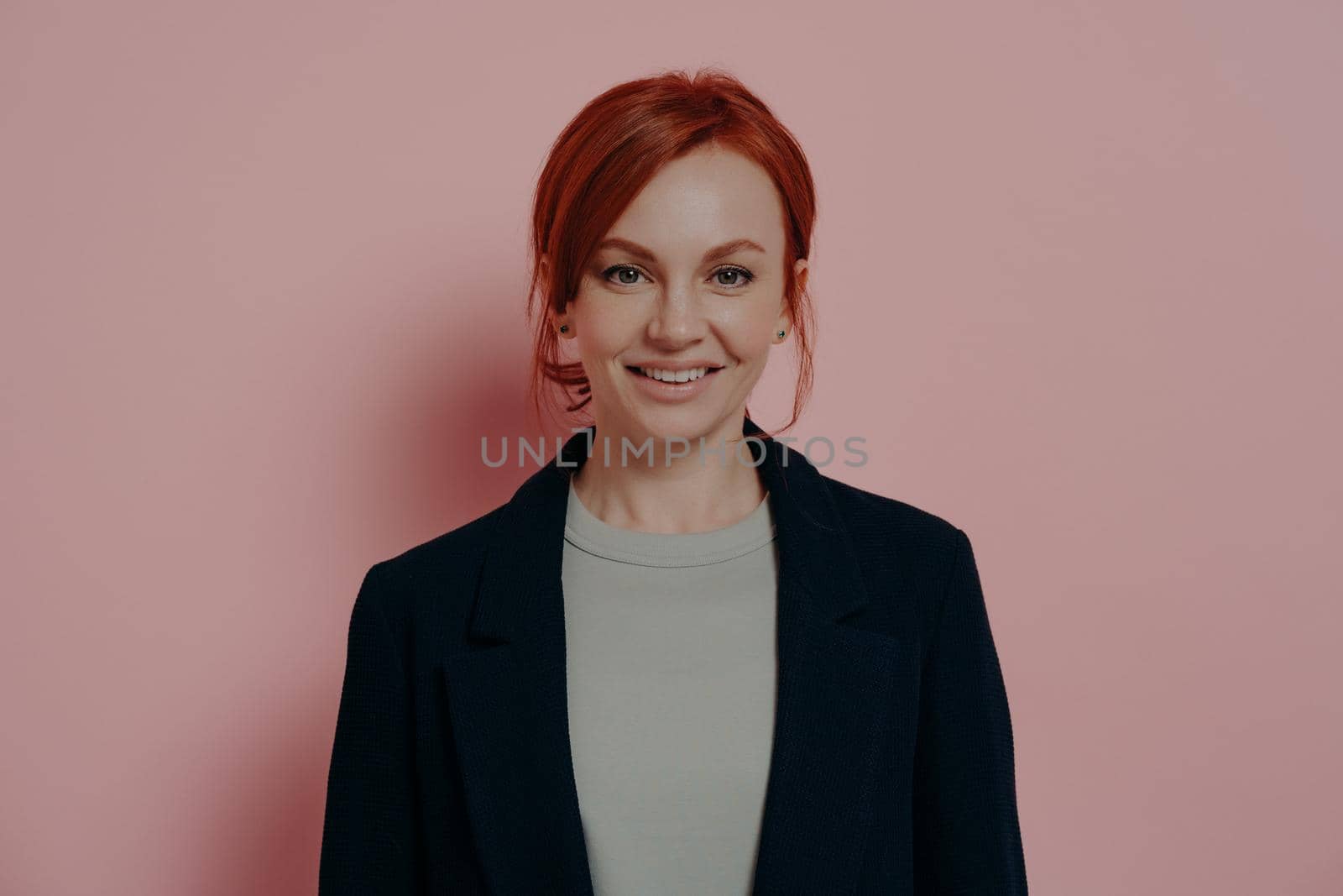 Young good-looking business woman with red hair looking positively at camera, isolated on pink by vkstock