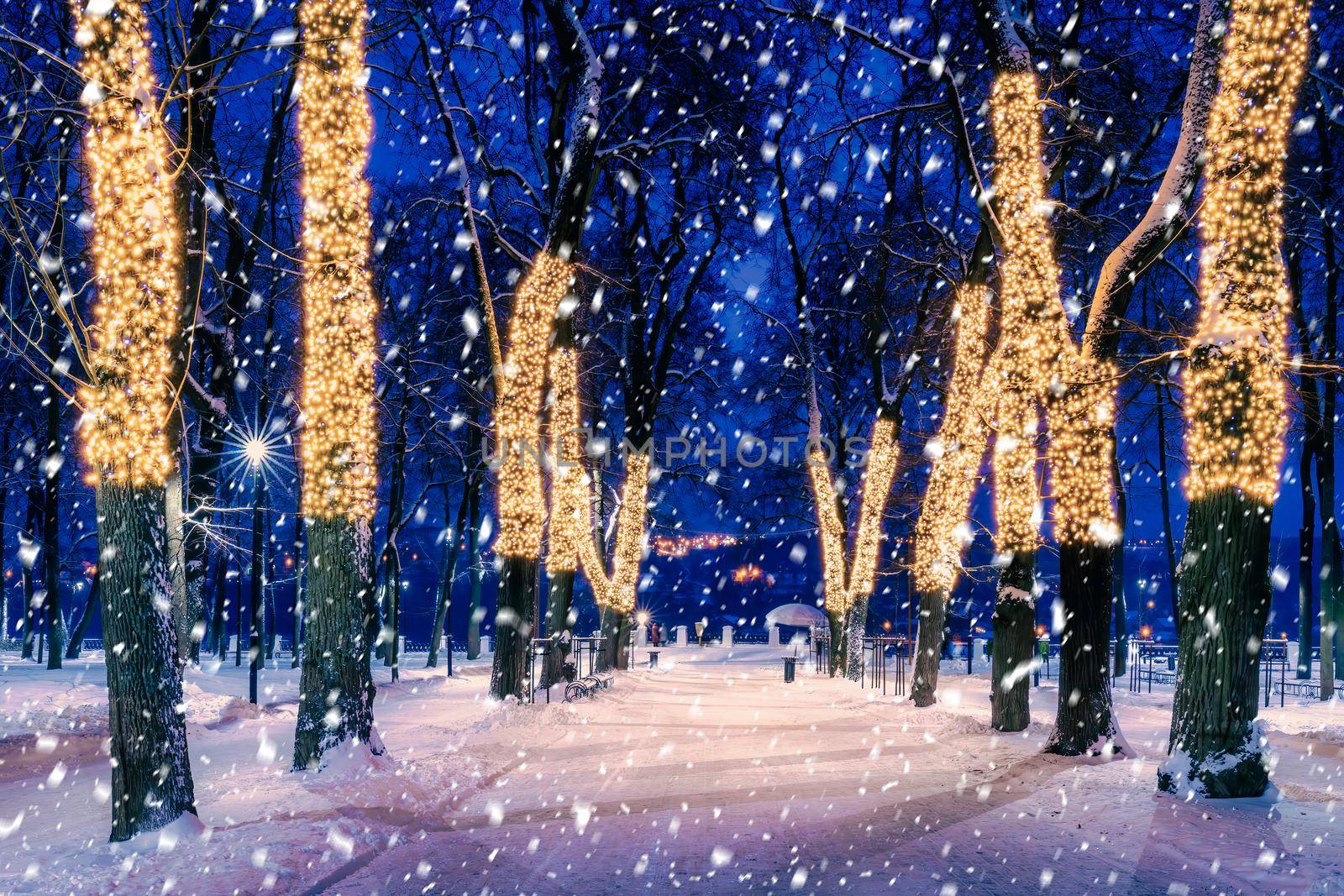 Snowfall in a winter park at night with christmas decorations, lights, pavement covered with snow and trees with garlands.