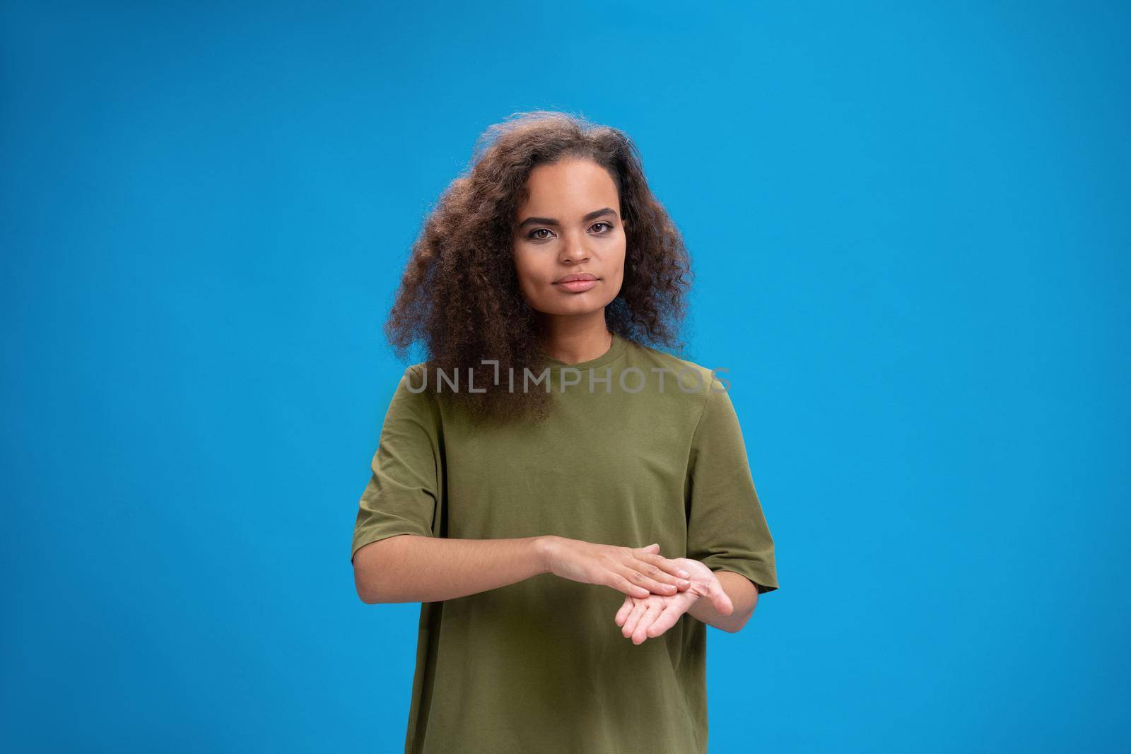 Deaf African American girl shows hand gesture using sign language. Young woman positively looking at camera wearing in an olive t-shirt. Isolated on blue background. Beauty concept. Skin care, hand cream.