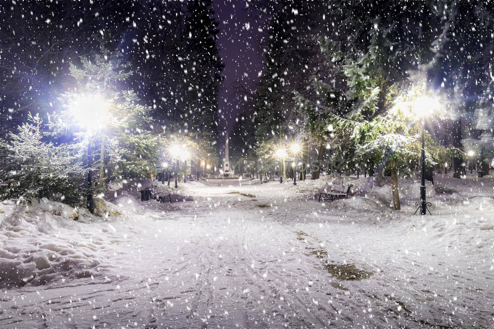 Snowfall in a winter park at night with christmas decorations, lights and  pavement covered with snow. Falling snow.