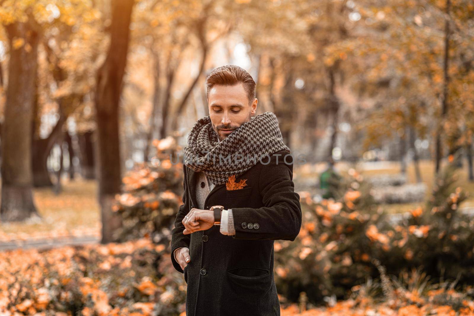 Waiting for his date handsome man looking at his arm watches wearing dark blue coat and scarf. Young freelancer man stands on the street in an autumn coat looking at time on his watches by LipikStockMedia