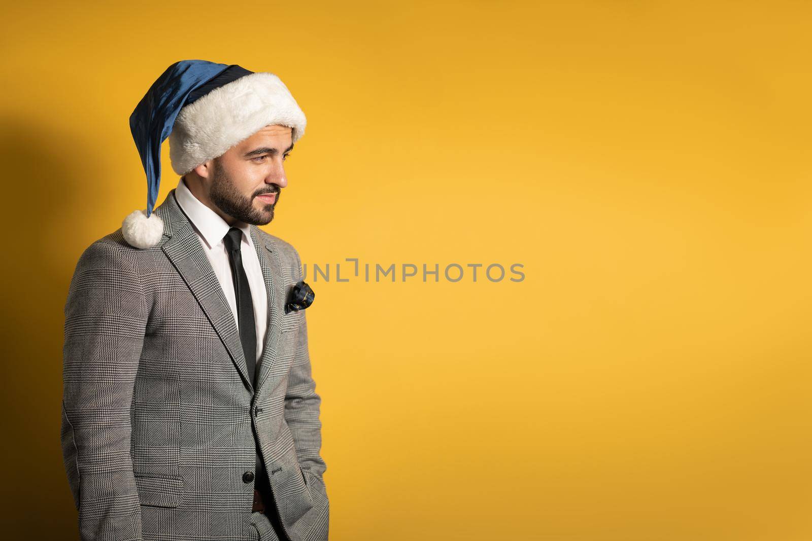 Stylish thoughtful handsome man wearing blue Santa Claus hat and suit, looking at camera isolated on yellow background. Positive emotions, feelings seasonal holidays.