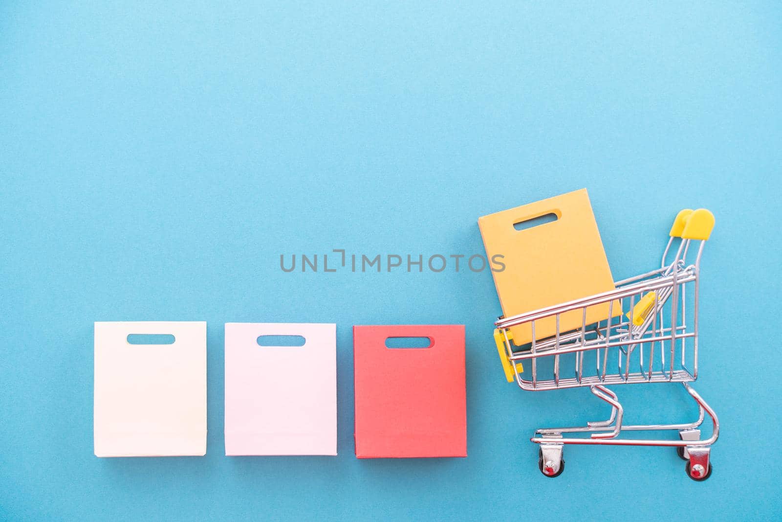 Abstract design element, annual sale, shopping season concept, mini yellow cart with colorful paper bag on pastel blue background, top view, flat lay