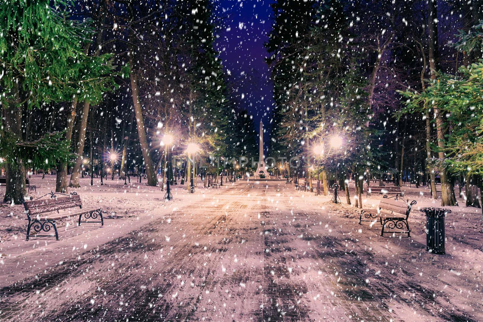 Snowfall in a winter park at night with christmas decorations, lights and  pavement covered with snow. Falling snow.