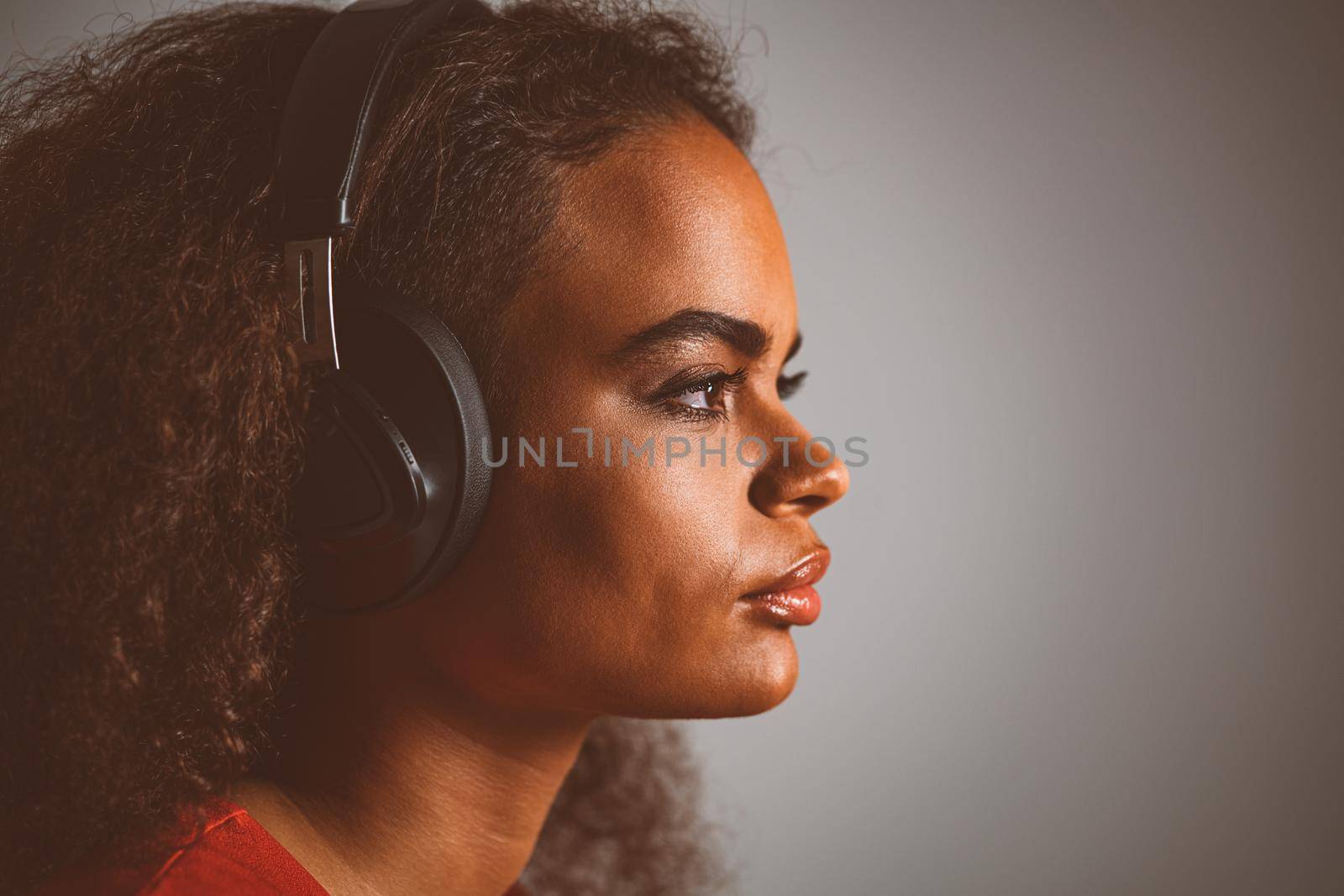 Catching a vibe by music young African-American girl looking up away sideways listening music wearing headphones, isolated on grey background, emotionally move, have fun. Close up. 