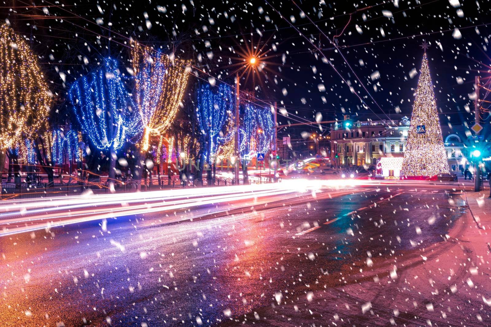 Night city with Christmas decorations, spruce and traces of headlights of moving cars, reflected in the wet road in a snowfall. by Eugene_Yemelyanov