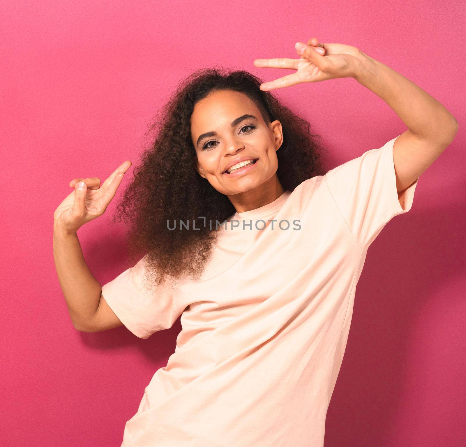 Young African American girl with Afro hair dancing gesturing peace with hands lifted up looking positively at camera wearing peachy t-shirt isolated on pink background. Beauty concept. Square footage.
