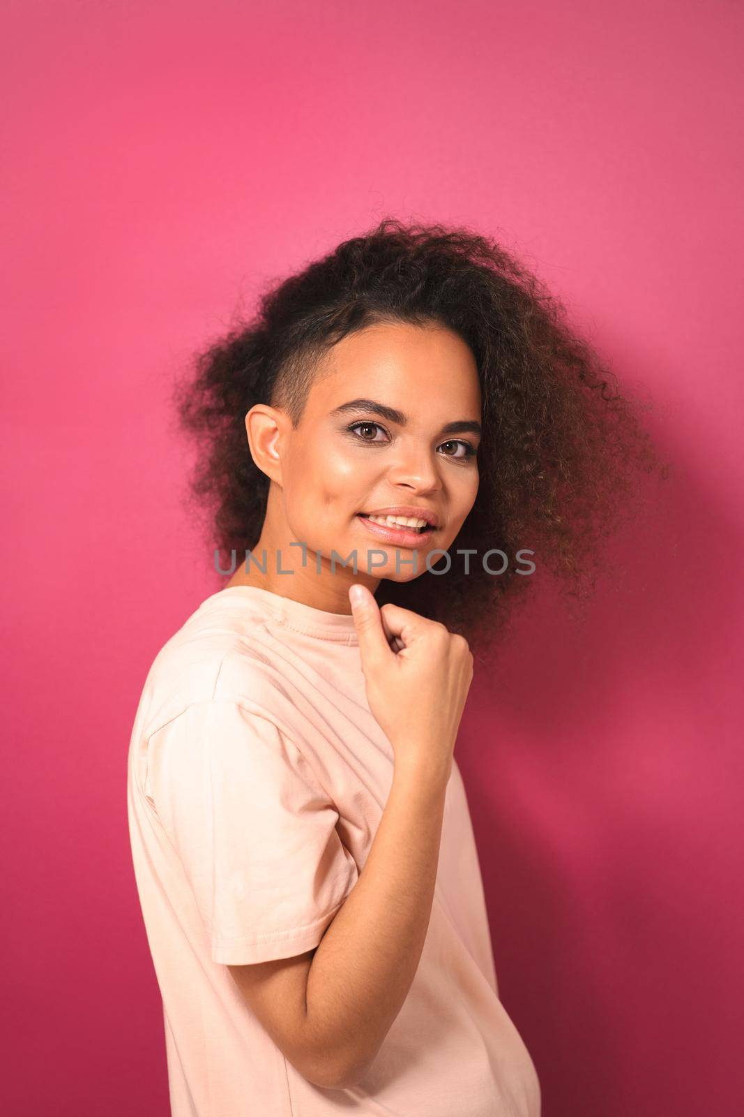 Charming passionate young African American girl with Afro hair looking positively at camera with one arm folded wearing peachy t-shirt isolated on pink background. Beauty concept by LipikStockMedia