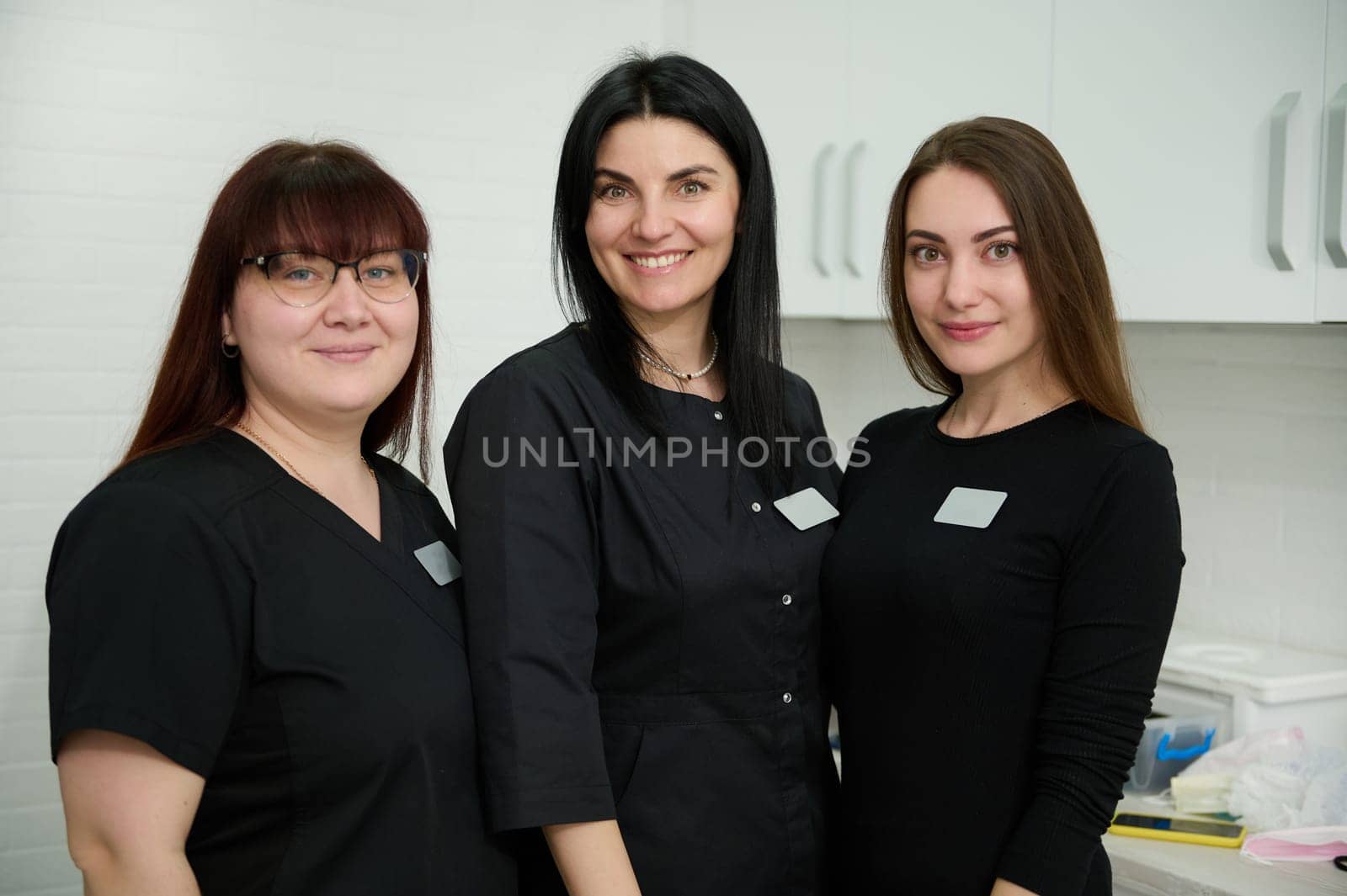 Confident portrait of beautiful diverse women, experienced team of female dentists doctors, dental hygienists in stylish black medical uniform, smiling looking at camera, standing in dentistry office