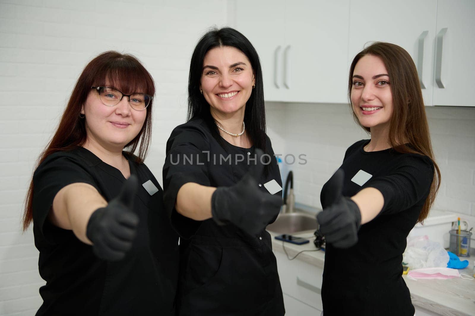 Confident team of female dentists doctors orthodontists in black medical uniform, smiling broadly looking at camera, gesturing with thumbs up, showing approval hands sign, in a dental office interior