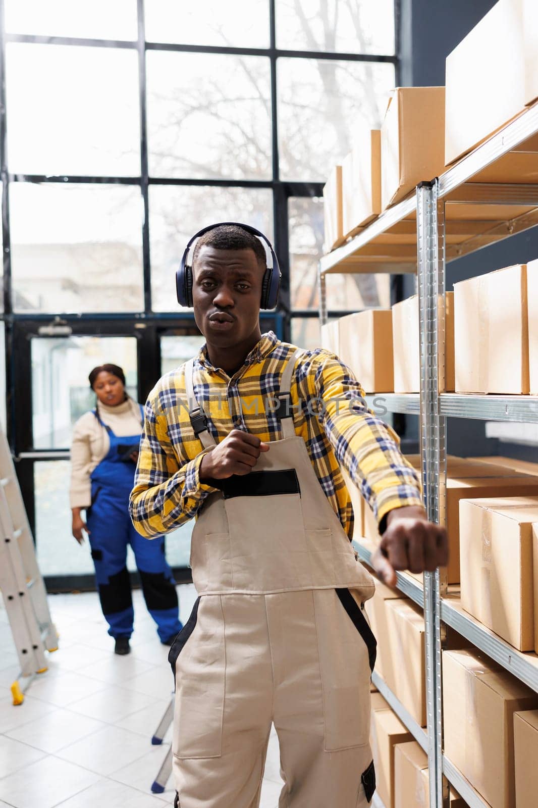 Warehouse package handler dancing at work and looking at camera. African american storehouse worker listening to playlist in headphones, singing along and making arms movements portrait