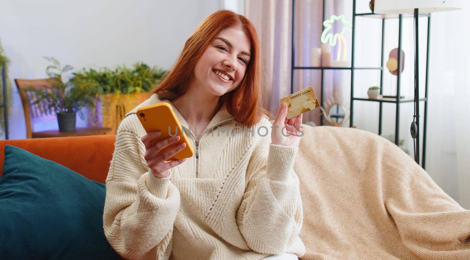 Young redhead woman using credit bank card and smartphone while transferring money, purchases online shopping cashless, order food delivery at home apartment indoors. Happy girl sits in room on couch