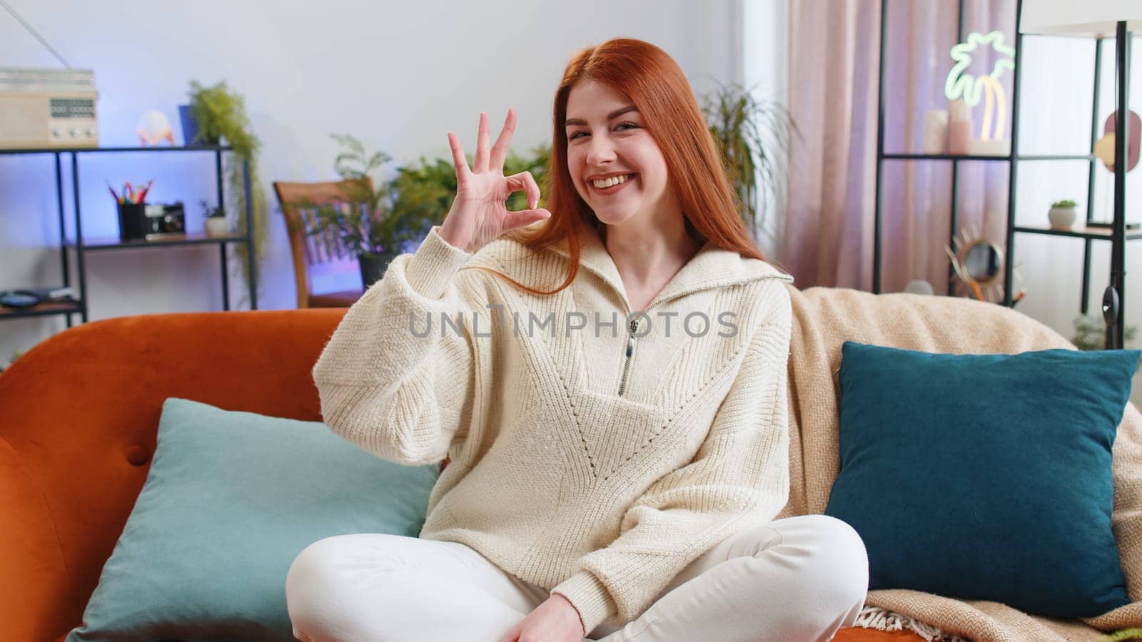 Okay gesture. Happy cheerful woman looking approvingly at camera showing, positive like sign, approve something good in living room sitting on sofa. Redhead girl at modern home apartment. Lifestyles