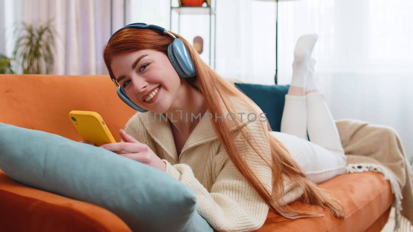 Happy overjoyed young woman in wireless headphones relaxing lying on couch at home apartment choosing listening favorite energetic disco dancing music. People weekend daytime leisure activities