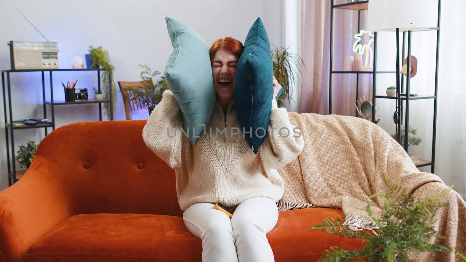 Repair work at neighbours. Irritated woman girl relaxing on couch cover ears with pillows annoyed by noisy neighbors suffer from headache wish silence. Thin walls at home flat without sound insulation