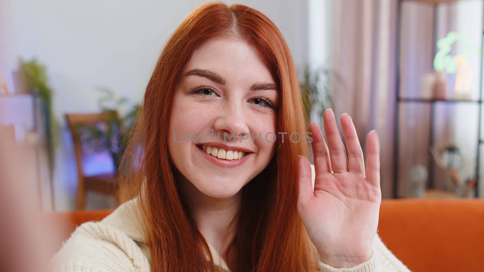 POV of young woman making online selfie video call talking waving hand hello, looking at camera at home apartment indoors. Communication, meeting. Redhead girl blogger making smartphone conversation
