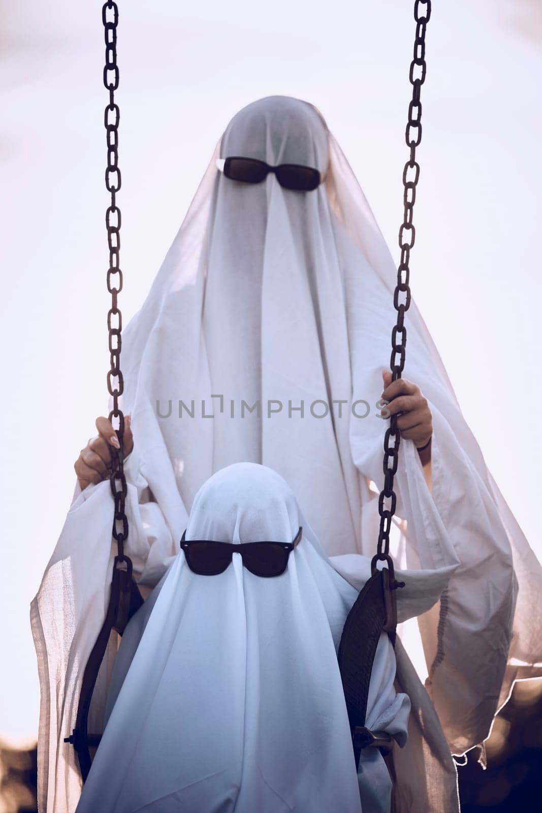 Halloween, ghost and family wearing white sheet and sunglasses costume at an outdoor park with child on a swing at playground. Horror, scary and celebrate fun holiday with creative woman and kid.