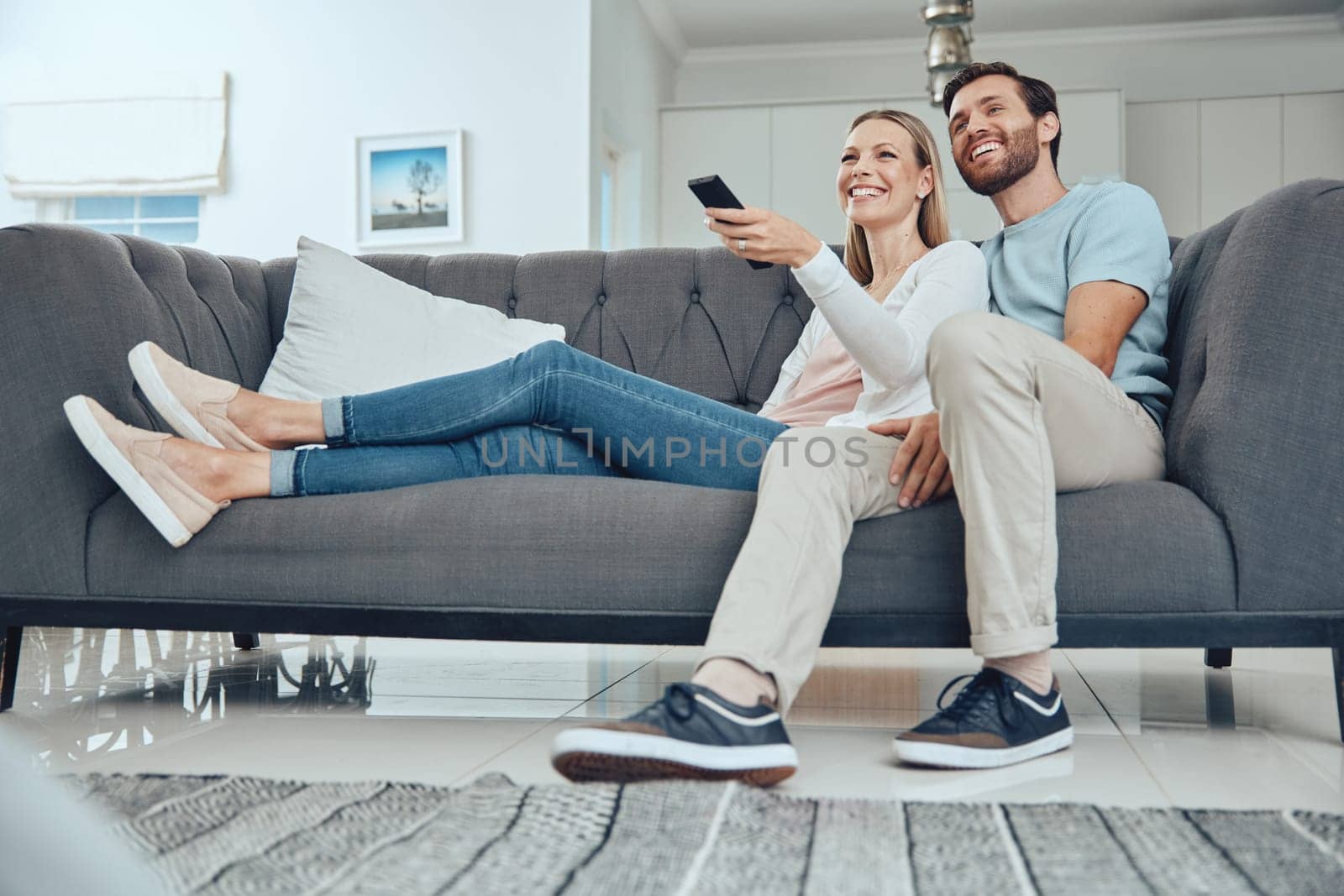 Couple, sofa and relax with tv remote, smile and happiness in home living room for bonding with love. Happy couple, couch and comedy on television, streaming or movie while laughing together in house by YuriArcurs