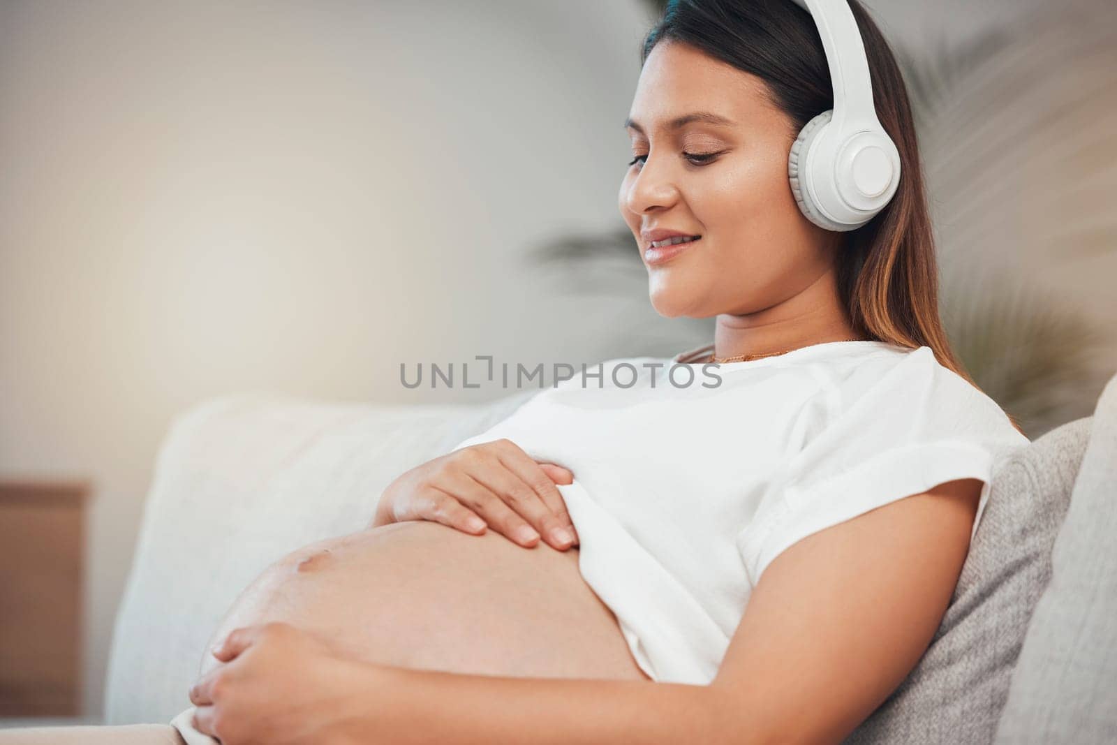 Woman, pregnant and relax while listening to calm music on sofa in healthy zen, wellness or meditation at home. Pregnant woman smile in relaxation holding tummy with headphones on living room couch by YuriArcurs