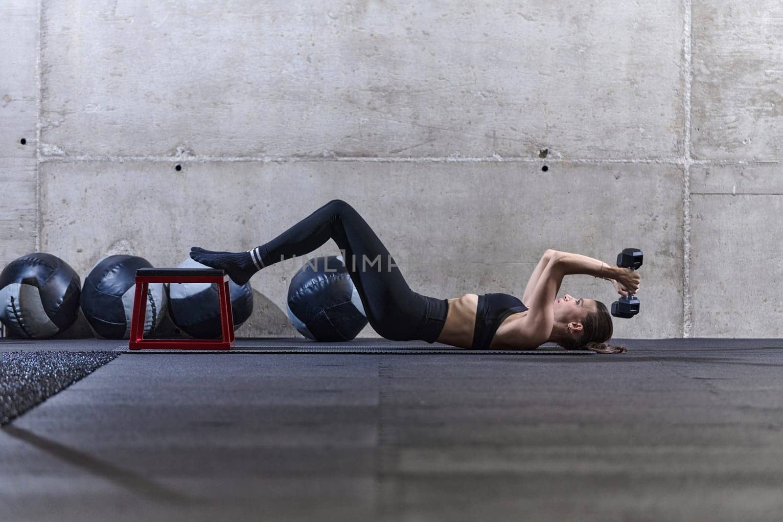 A fit woman is lying on the gym floor, performing arm exercises with dumbbells and showcasing her dedication and strength