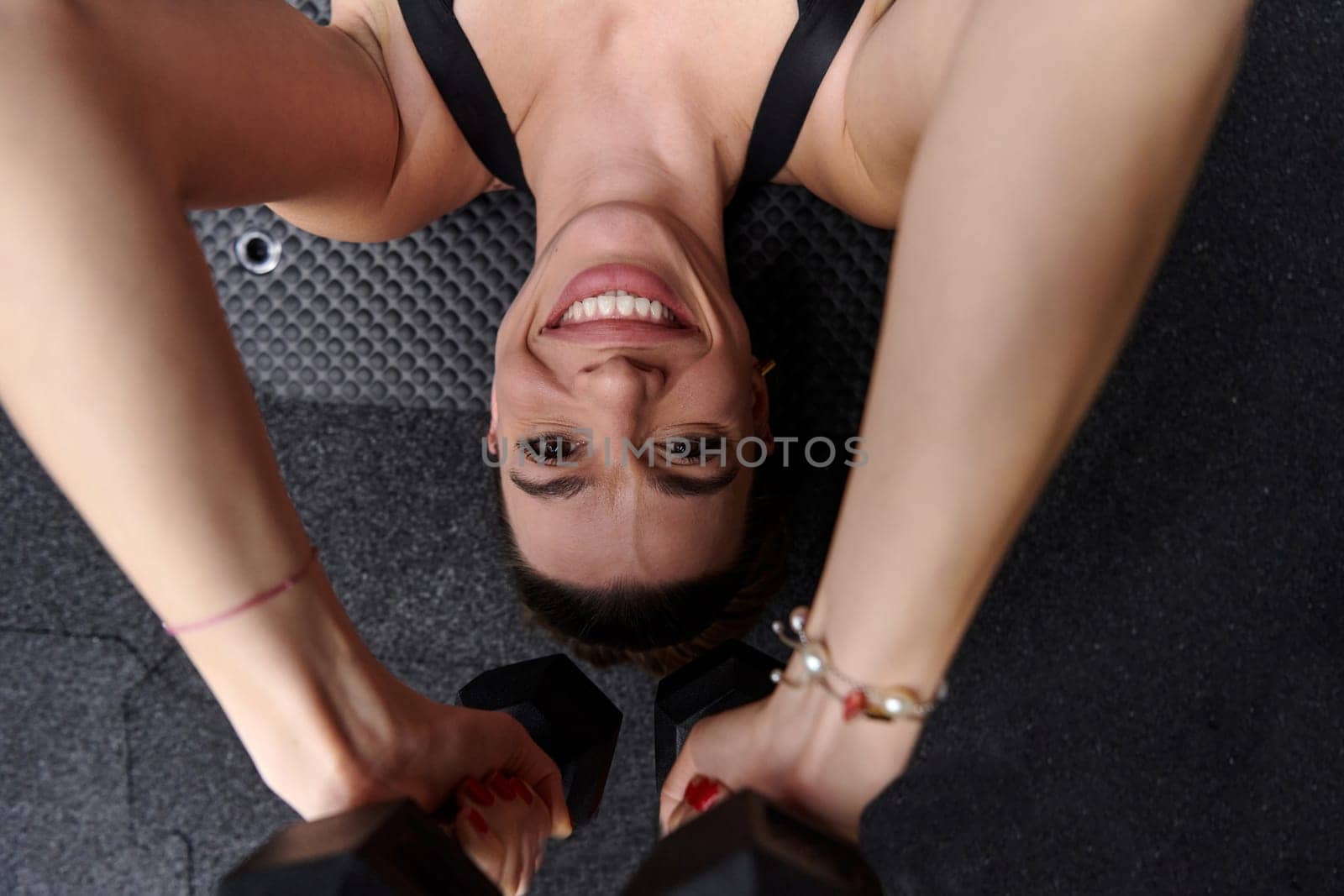 A fit woman is lying on the gym floor, performing arm exercises with dumbbells and showcasing her dedication and strength