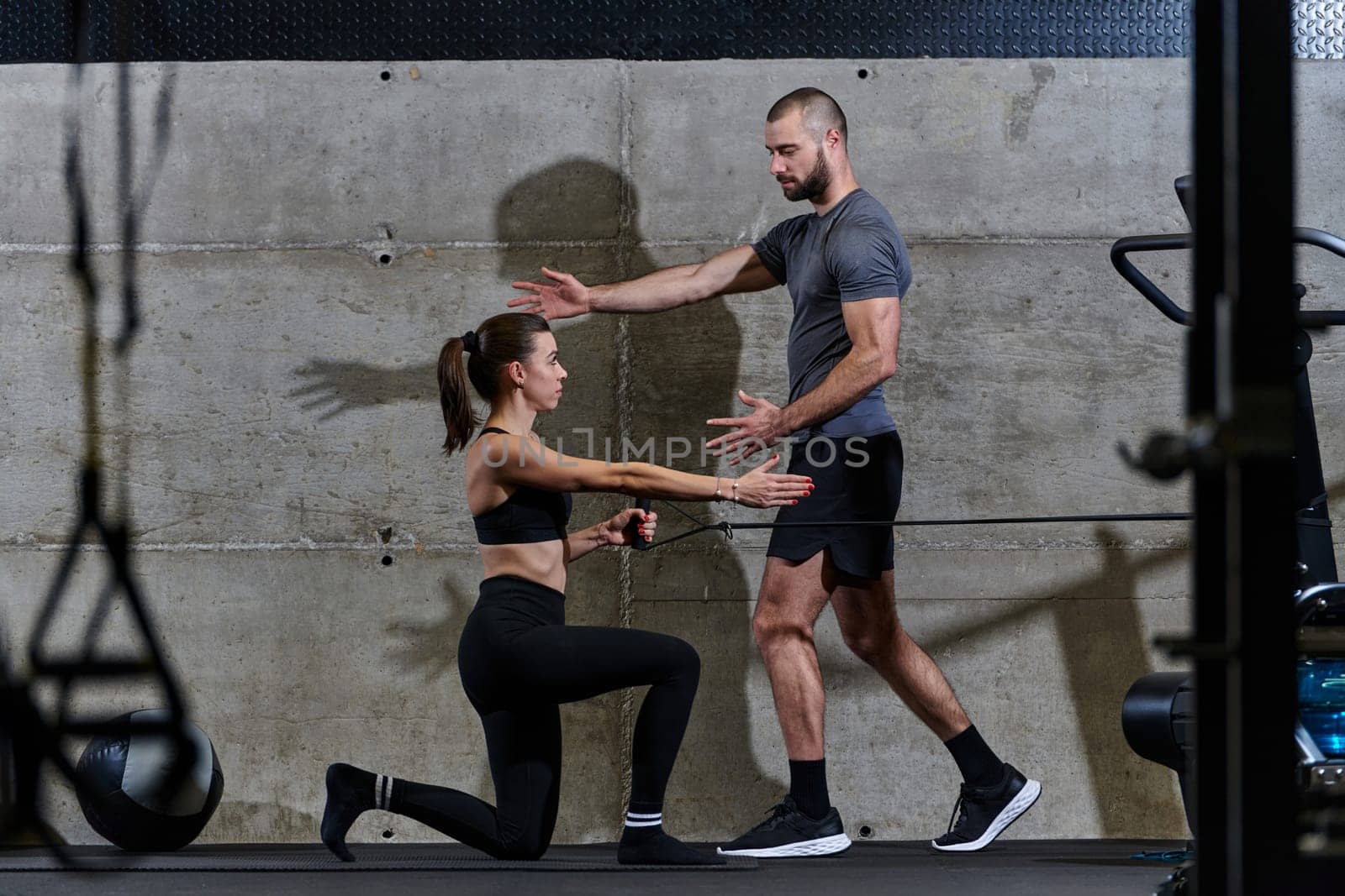 A muscular man assisting a fit woman in a modern gym as they engage in various body exercises and muscle stretches, showcasing their dedication to fitness and benefiting from teamwork and support.