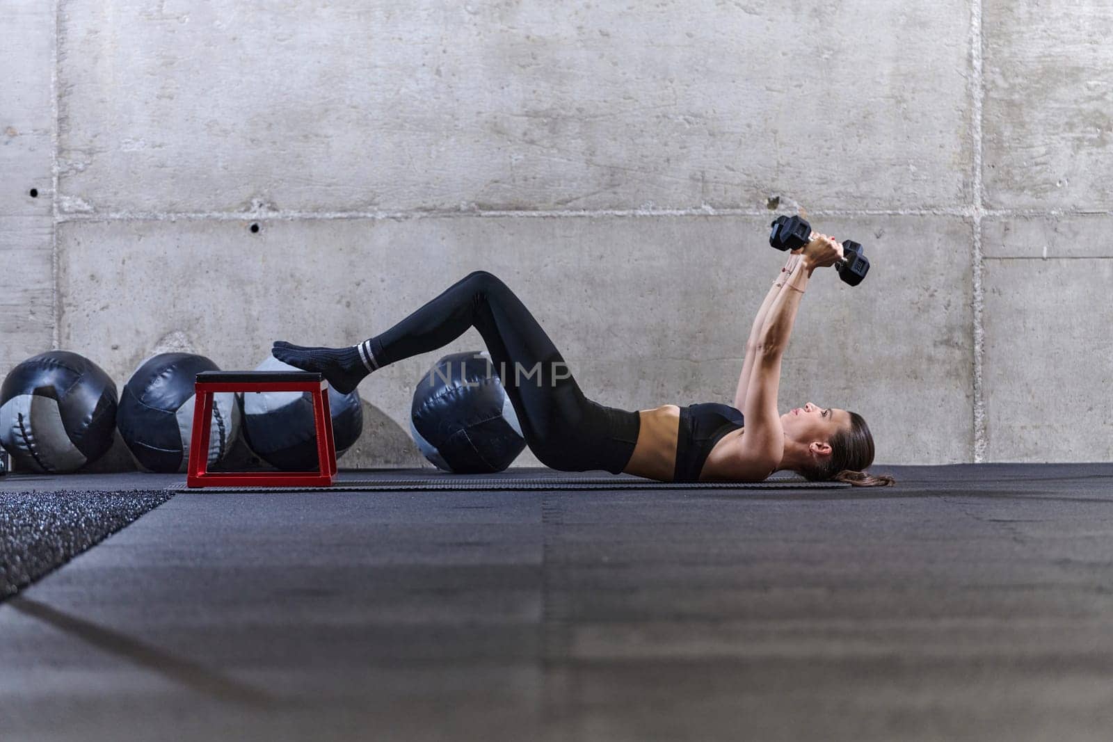 A fit woman is lying on the gym floor, performing arm exercises with dumbbells and showcasing her dedication and strength