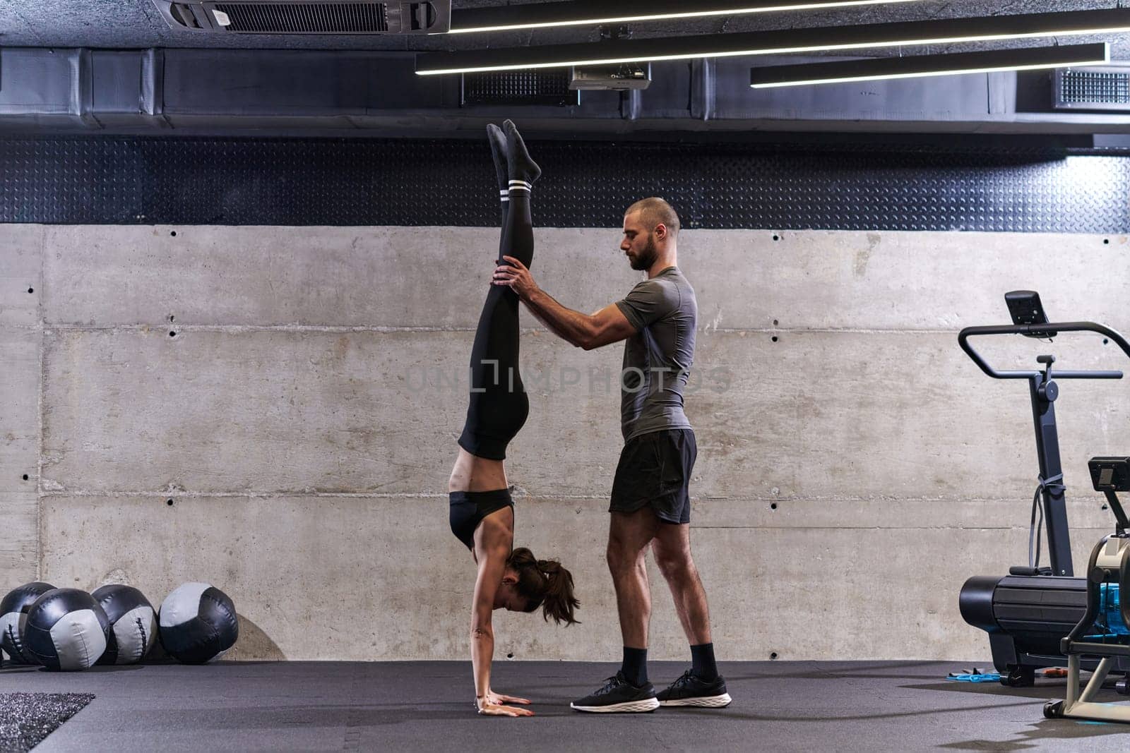 A muscular man assisting a fit woman in a modern gym as they engage in various body exercises and muscle stretches, showcasing their dedication to fitness and benefiting from teamwork and support by dotshock