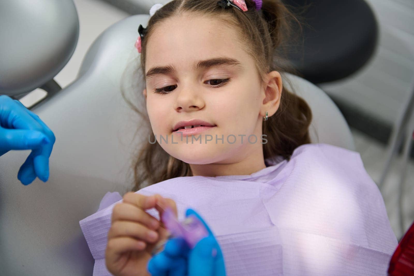 Close-up little kid girl in dentist's chair at dental check-up, listens to dentist how to use a dental floss for cleaning teeth. Dental care and oral hygiene concept. Pediatric dentistry concept