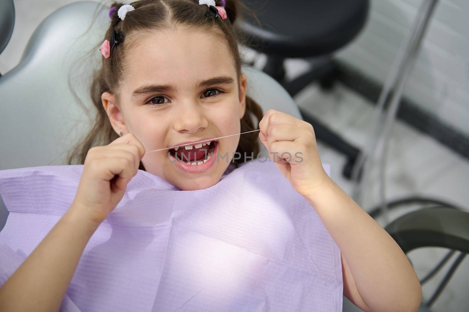 Close-up portrait of beautiful little girl using dental floss, brushing her teeth, sitting in the dentist's chair and smiling cutely, looking at the camera. Oral hygiene concept for caries prevention