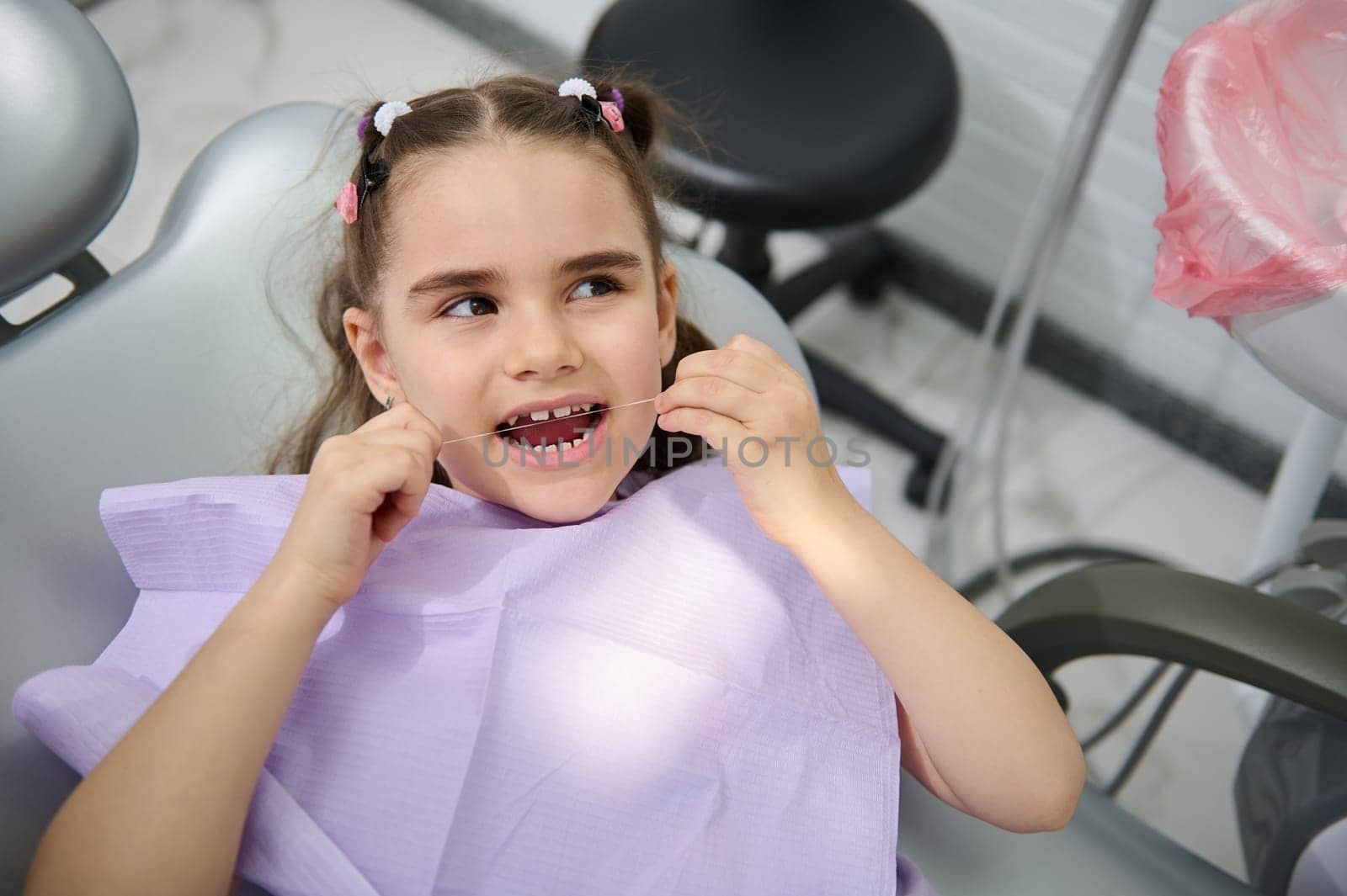 Smiling child girl, a patient in pediatric dentistry clinic, cleaning her teeth with a dental floss. Oral care and hygiene for prevention caries of baby teeth. Dental practice. Healthcare and medicine