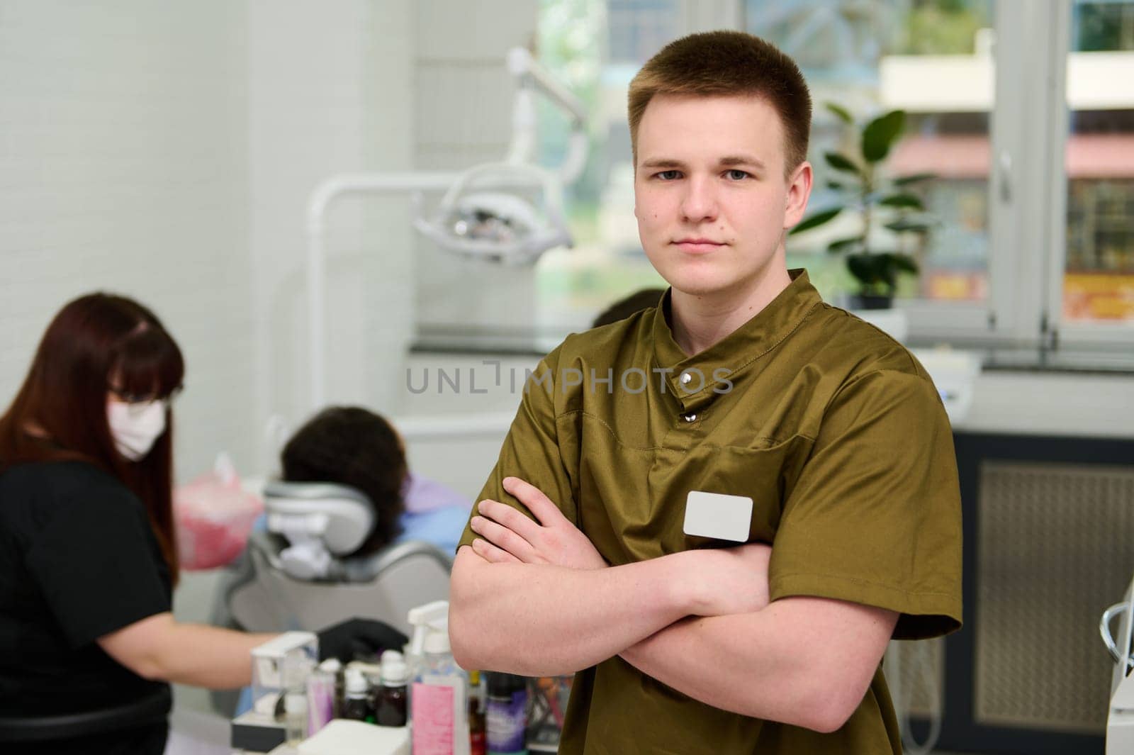 Confident portrait of a Caucasian young happy male dentist orthodontist in medical uniform, standing with arms folded in the dental office in modern dentistry clinic, smiling looking at camera